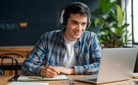 Joven con audífonos tomando notas frente a su computadora portátil