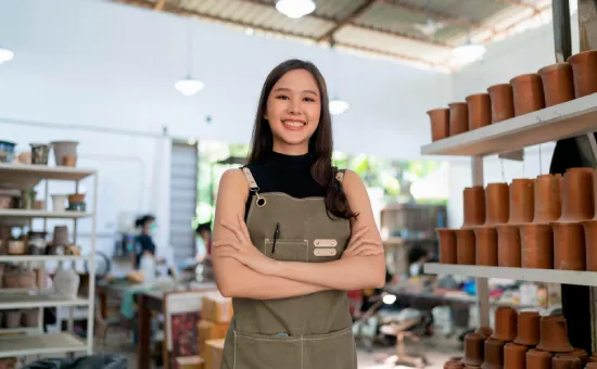 Cómo la universidad puede ayudarte a crear y hacer crecer tu emprendimiento Joven sonriente con delabel en taller de cerámica rodeada de estantes con piezas de arcilla